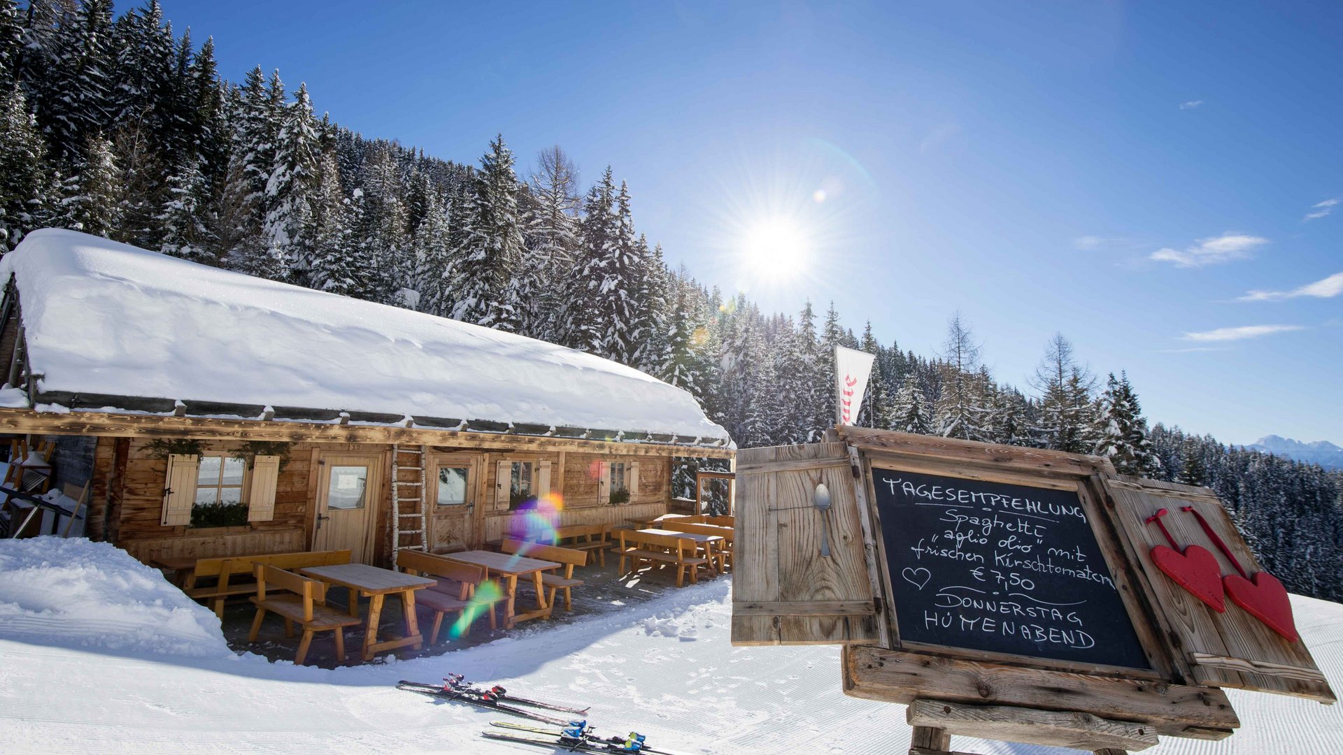 Rund um Meransen: Hütten und Almen Auf dem Bild ist eine verschneite Berghütte mit Holzbänken und Tischen vor der Hütte zu sehen; daneben steht eine Tafel, die die Tagesempfehlung anzeigt, während im Hintergrund verschneite Nadelbäume und ein strahlend blauer Himmel zu sehen sind.