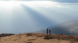 De mooiste wandelroutes in de Dolomieten De afbeelding toont twee wandelaars die op een met gras begroeide heuvel staan, omgeven door een uitgestrekt bergachtig landschap. Stralen zonlicht breken dramatisch door de wolken en verlichten de mist in de verte, wat een sfeervolle en vredige scène creëert.