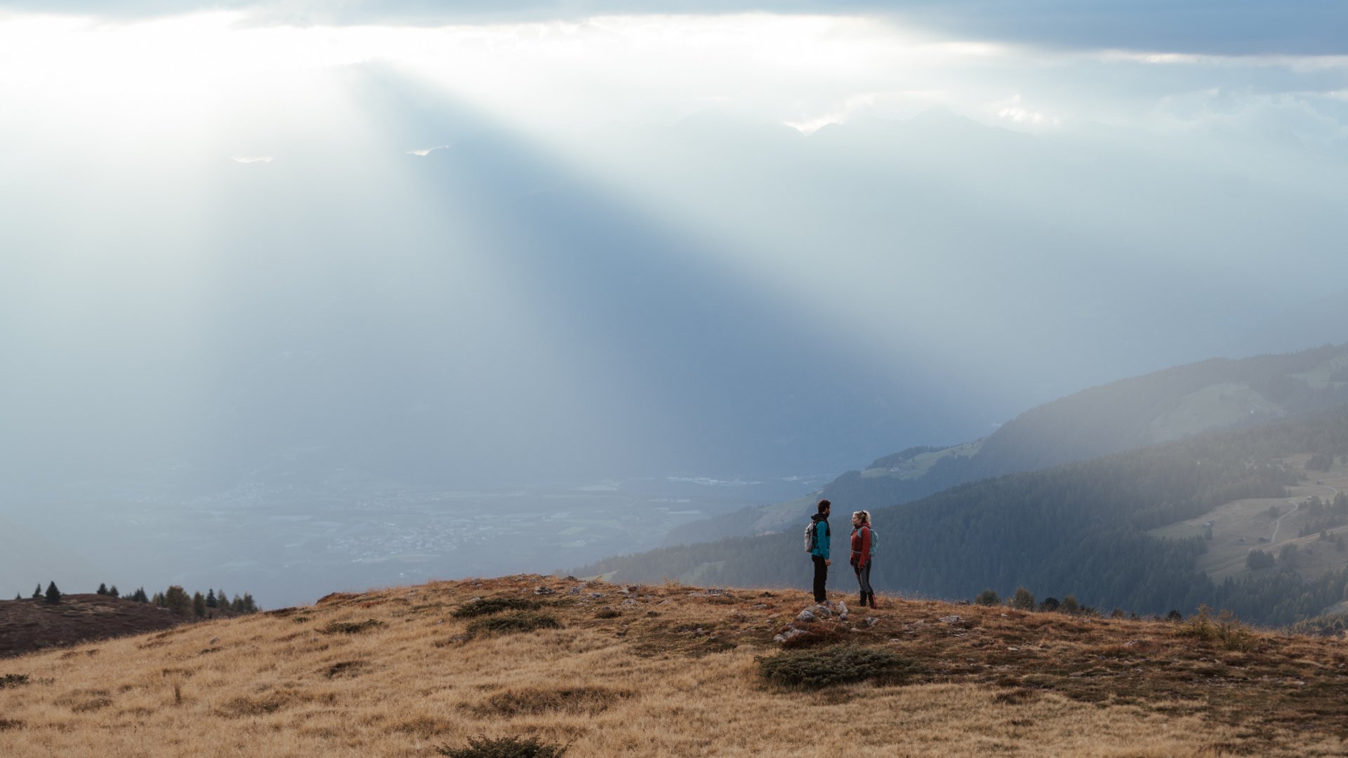 De mooiste wandelroutes in de Dolomieten De afbeelding toont twee wandelaars die op een met gras begroeide heuvel staan, omgeven door een uitgestrekt bergachtig landschap. Stralen zonlicht breken dramatisch door de wolken en verlichten de mist in de verte, wat een sfeervolle en vredige scène creëert.