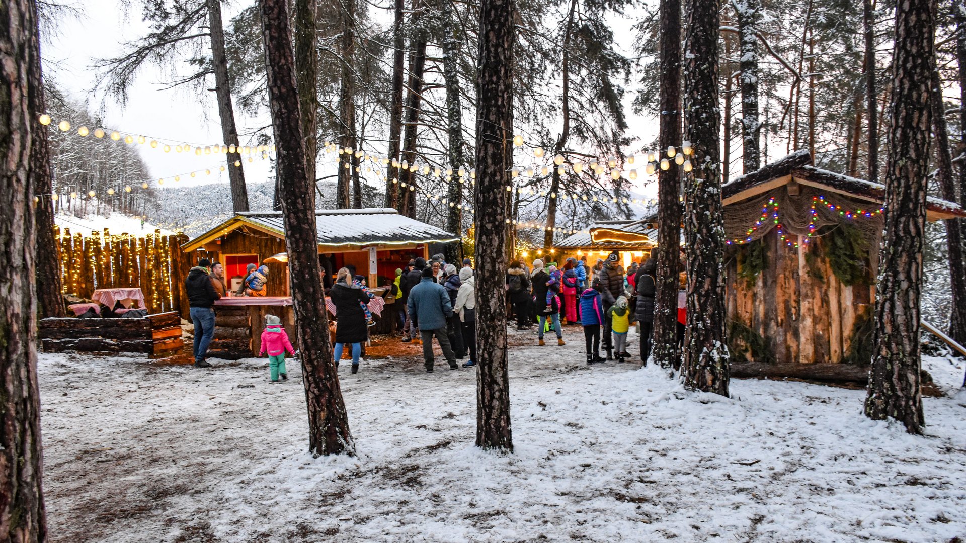 Advent in Terenten The picture shows a Christmas market in the forest with small wooden huts decorated with string lights. People in winter clothing stand in front of the huts, and the ground is covered with a light layer of snow.