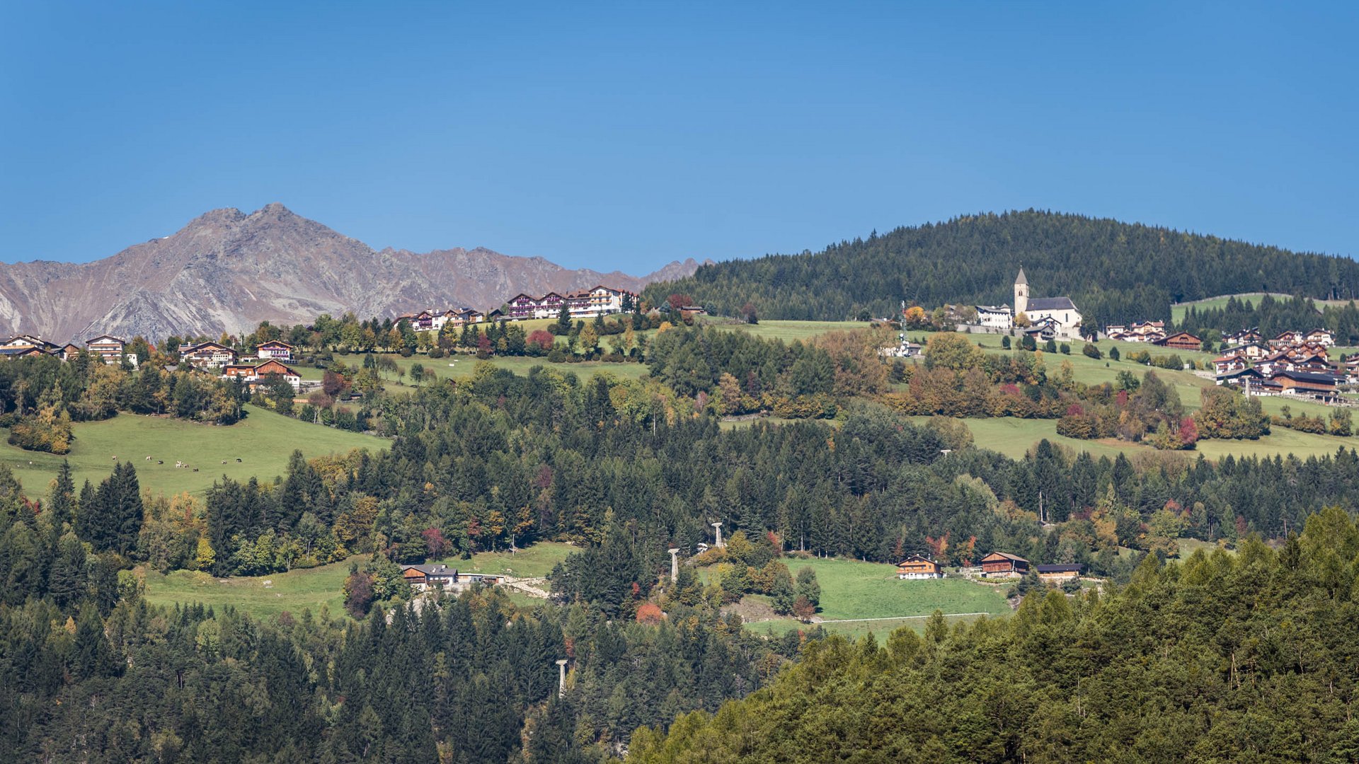 The mountain village of Meransen The picture shows a picturesque landscape with a village spreading over green hills. In the foreground, dense forests are visible, while in the background high mountains rise under a clear blue sky. The houses and a church of the village are scattered among the green hills.