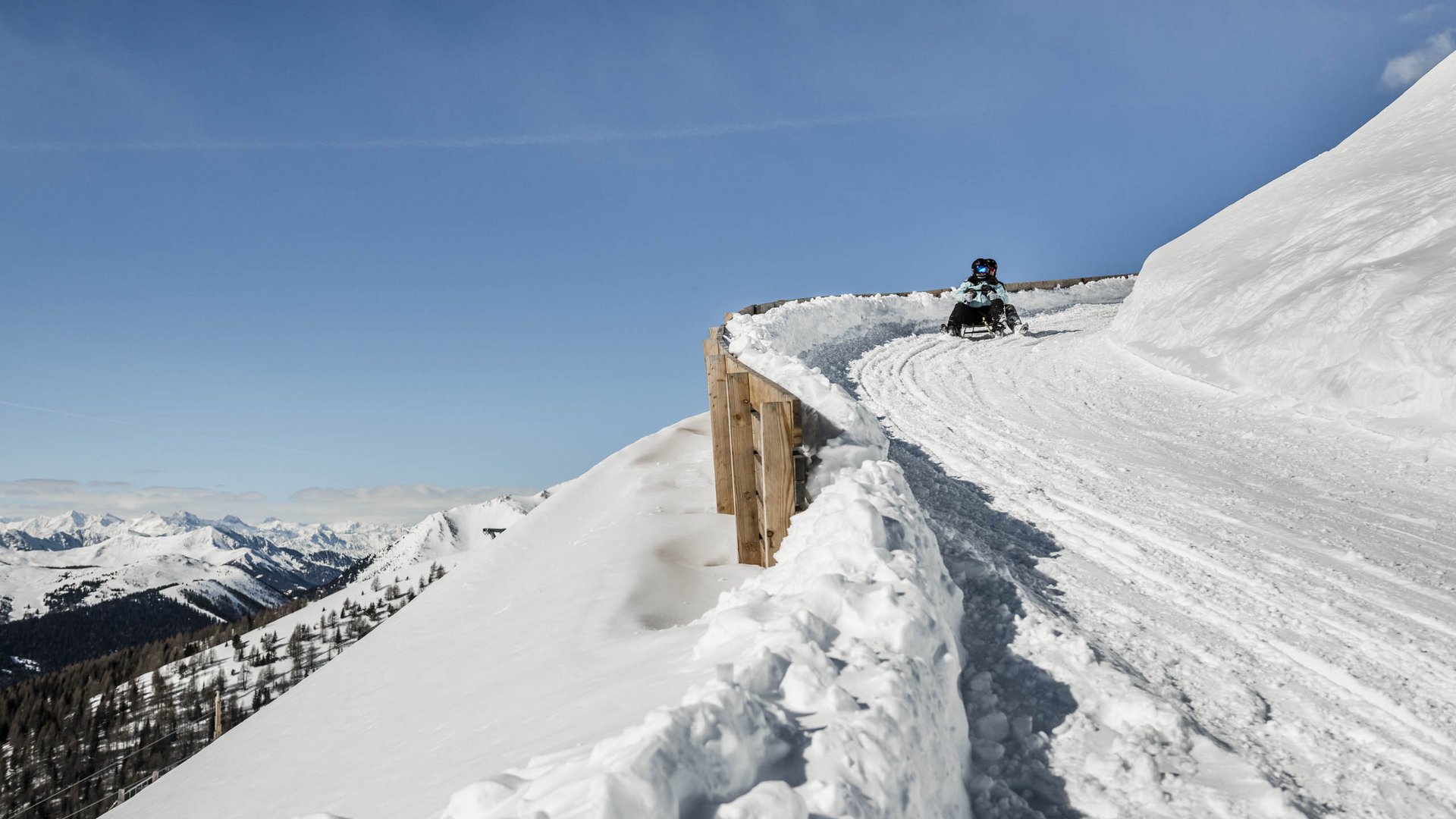 Rodeln in Südtirol Person rodelnd auf einer schneebedeckten Bergpiste unter blauem Himmel