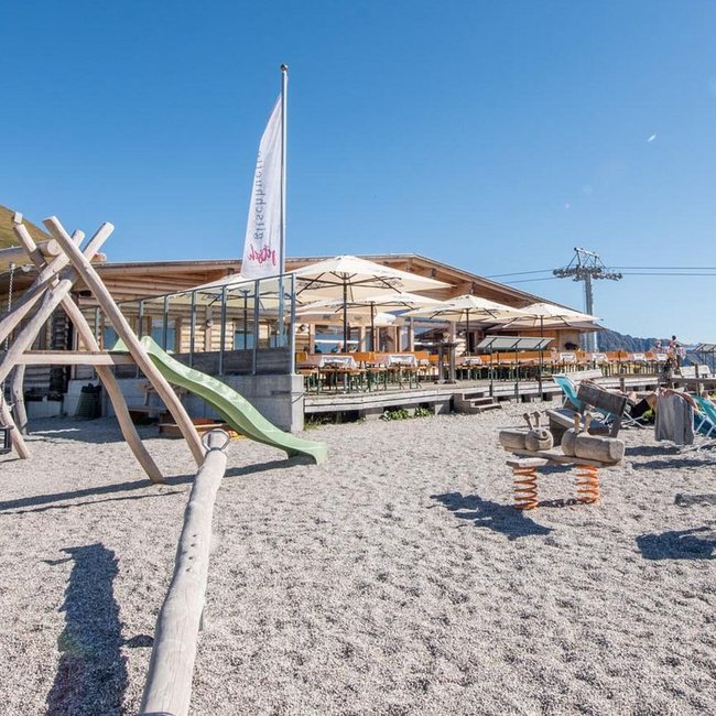Gitschhütte Eine Berghütte mit Terrasse und Sonnenschirmen steht auf einer Wiese und daneben ein Spielplatz und Liegestühle. Im Hintergrund sind Berge und ein Sessellift zu sehen.