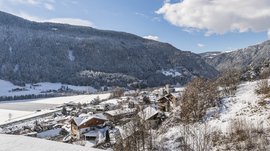 Vintl im Pustertal Das Bild zeigt ein verschneites Dorf, das in einem Tal zwischen dicht bewaldeten, schneebedeckten Bergen liegt. Im Vordergrund sieht man einige Häuser mit Schnee auf den Dächern, und im Hintergrund erhebt sich ein großer, bewaldeter Berg unter einem blauen Himmel mit wenigen Wolken.