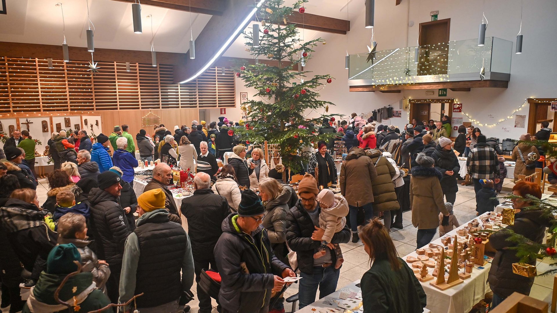 Tearna Advent Menschen auf Weihnachtsmarkt mit Tannenbaum und Ständen in einer Halle
