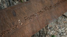 Der Schöpfungsweg in Rodeneck Das Bild zeigt eine rostige Metalltafel, auf der der Schriftzug "Tag 5 Fische und Vögel" eingestanzt ist. Der Hintergrund besteht aus natürlichen Elementen wie Erde, Steinen und Baumrinde.