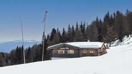 Hiking in Spinges, South Tyrol – no map needed! The picture shows the "Anratter Hütte," a wooden hut on a snow-covered slope, surrounded by coniferous trees. In the background, snow-covered mountain peaks are visible under a blue sky. A red flag flies on one of the flagpoles in front of the hut.