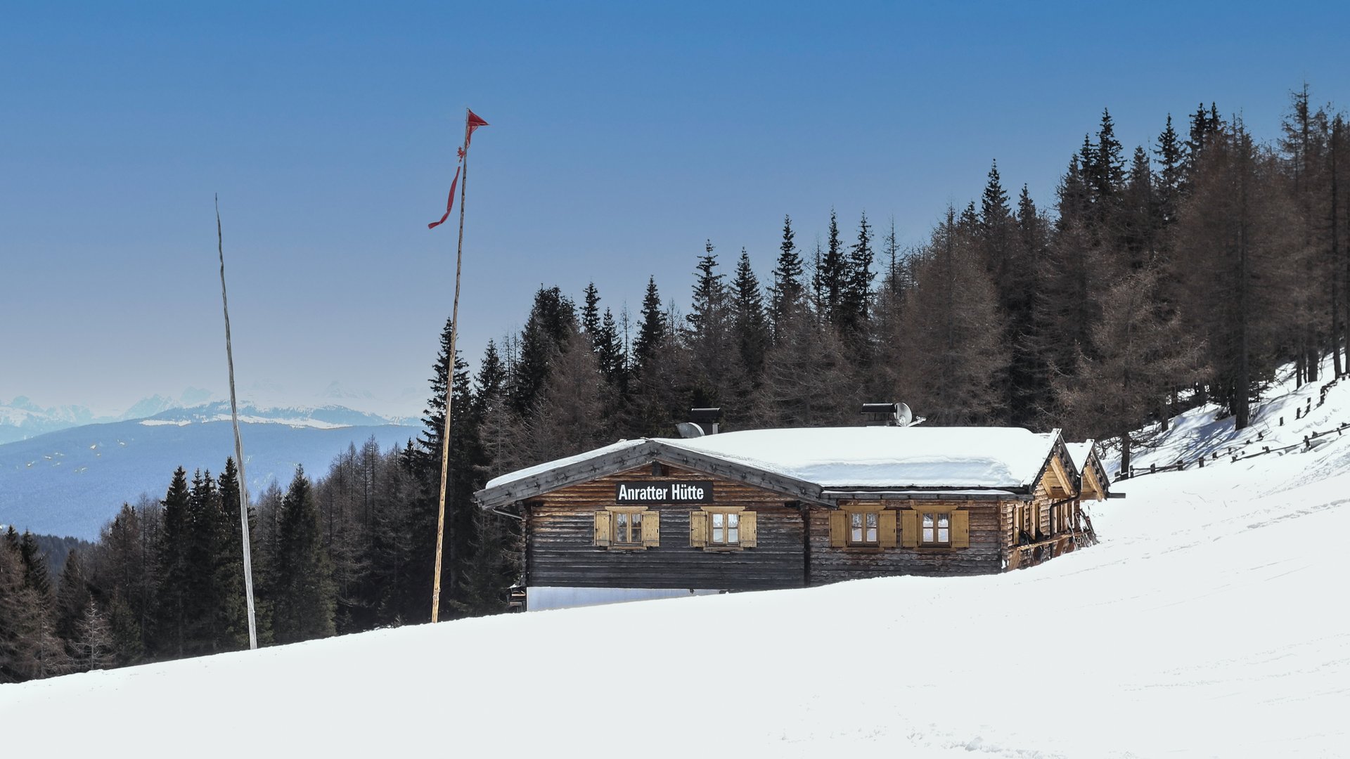 Hiking in Spinges, South Tyrol – no map needed! The picture shows the "Anratter Hütte," a wooden hut on a snow-covered slope, surrounded by coniferous trees. In the background, snow-covered mountain peaks are visible under a blue sky. A red flag flies on one of the flagpoles in front of the hut.