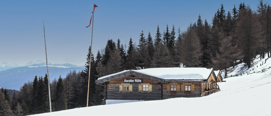 The Gitschberg webcams The picture shows the "Anratter Hütte," a wooden hut on a snow-covered slope, surrounded by coniferous trees. In the background, snow-covered mountain peaks are visible under a blue sky. A red flag flies on one of the flagpoles in front of the hut.