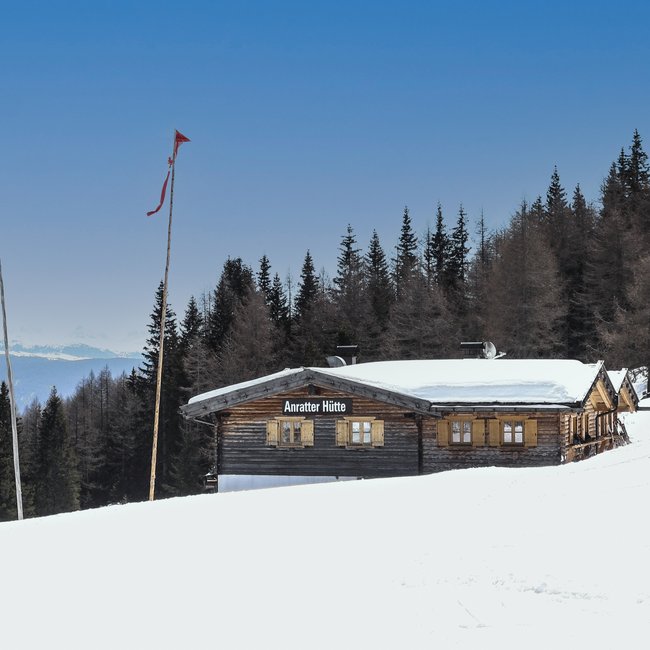 Anratterhütte Das Bild zeigt die "Anratter Hütte," eine Holzhütte auf einem schneebedeckten Hang, umgeben von Nadelbäumen. Im Hintergrund sind verschneite Berggipfel unter einem blauen Himmel zu sehen. Eine rote Fahne weht an einer der Fahnenstangen vor der Hütte.