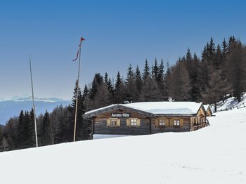 The Alpine hamlet of Spinges The picture shows the "Anratter Hütte", a wooden hut on a snow-covered slope, surrounded by conifers. Snow-covered mountain peaks can be seen in the background under a blue sky. A red flag flies on one of the flagpoles in front of the hut.