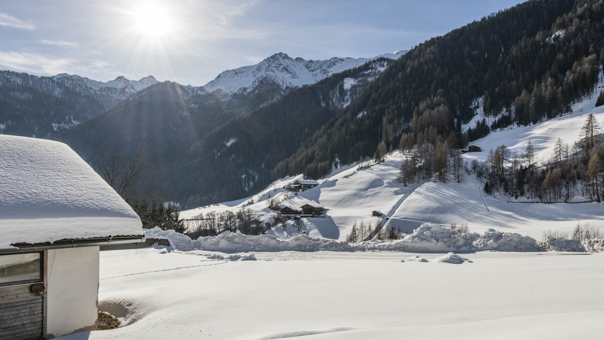 Pfunders: urig und idyllisch Das Bild zeigt eine schneebedeckte Landschaft mit Hügeln und Bergen im Hintergrund. Im Vordergrund ist ein Gebäude mit einem schneebedeckten Dach zu sehen, während die Sonne hell am Himmel scheint und über die verschneiten Hügel strahlt.