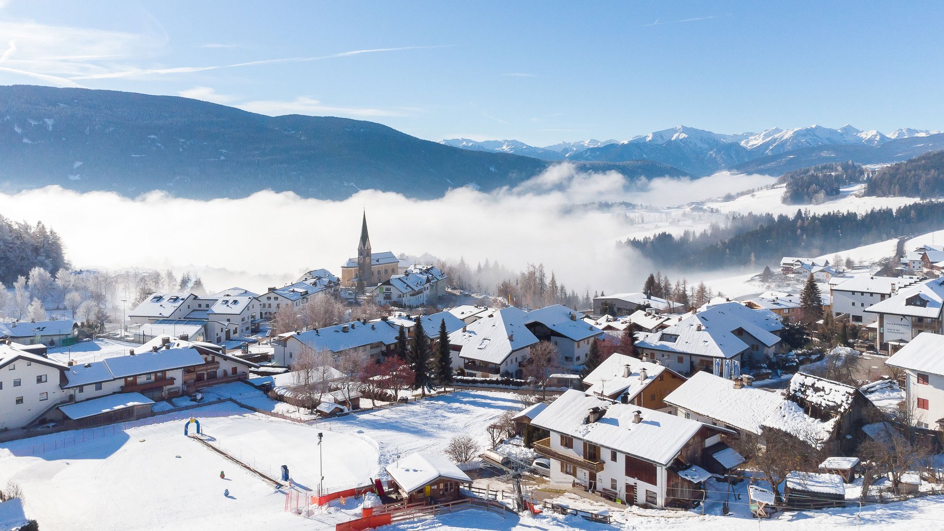 Skivakantie met kinderen in Zuid-Tirol/Terenten De afbeelding toont een met sneeuw bedekt dorp met huizen en een kerk te midden van een winterse berglandschap. Op de achtergrond trekken nevelslierten door het dal, terwijl de lucht helder en blauw is.
