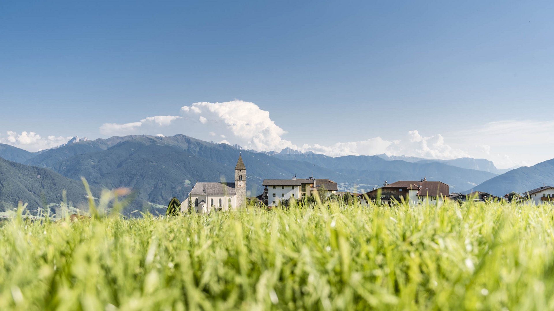 The mountain village of Meransen The picture shows a meadow with tall, green grass in the foreground. Behind it, there is a small village with a church and several houses. In the background, tall mountains can be seen under a clear, blue sky.