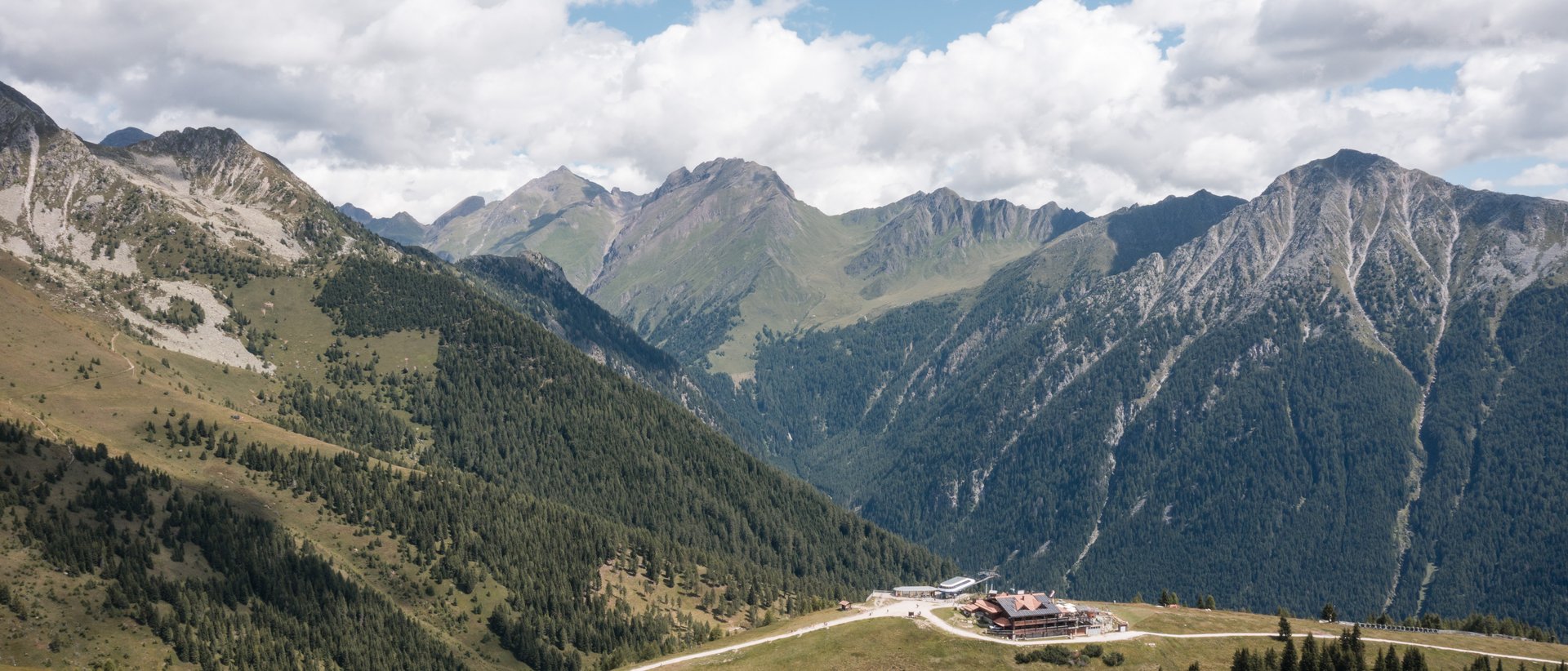 Die Almencard speziell für euch Berglandschaft mit Tannenwäldern, einem Gebäude und bewölktem Himmel