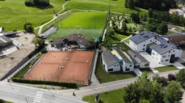 Het Aktivpark Terenten De afbeelding toont een luchtfoto van sportvoorzieningen in een landelijke omgeving. Te zien zijn twee tennisbanen, een groot voetbalveld en enkele gebouwen, waaronder stallen met paarden aan de linkerkant. De omgeving bestaat uit groene weiden en heuvels.