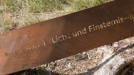 Der Schöpfungsweg in Rodeneck Das Bild zeigt eine rostfarbene Metalltafel im Gras. In die Tafel sind die Wörter „Tag 1 - Licht und Finsternis“ eingraviert.