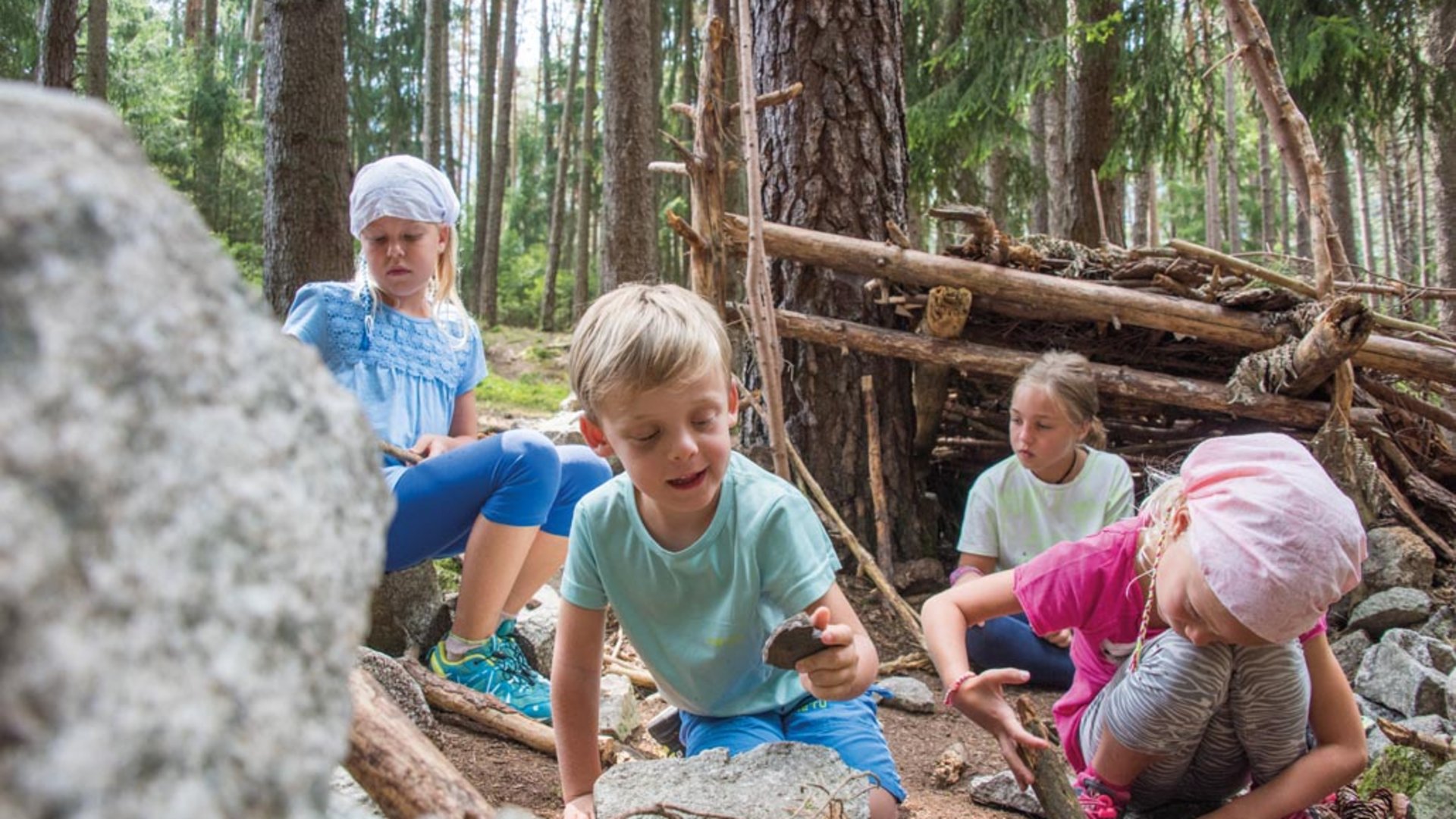 De Elfenweg bij Vintl De afbeelding toont vier kinderen die in een bos zitten en spelen met stenen en stokjes. Op de achtergrond staat een geïmproviseerde houten hut, gebouwd van takken. De kinderen lijken geconcentreerd te spelen met de natuurlijke materialen.