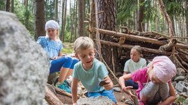 De Elfenweg bij Vintl De afbeelding toont vier kinderen die in een bos zitten en spelen met stenen en stokjes. Op de achtergrond staat een geïmproviseerde houten hut, gebouwd van takken. De kinderen lijken geconcentreerd te spelen met de natuurlijke materialen.