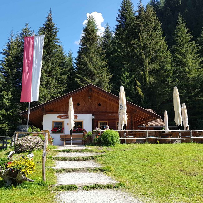 Kurzkofelhütte Een berghut met een terras waarop gesloten parasols staan, is omringd door een groen weiland en hoge sparren. Voor de hut wappert een grote rood-witte vlag, en bloemen bloeien in een decoratieve houten kar.