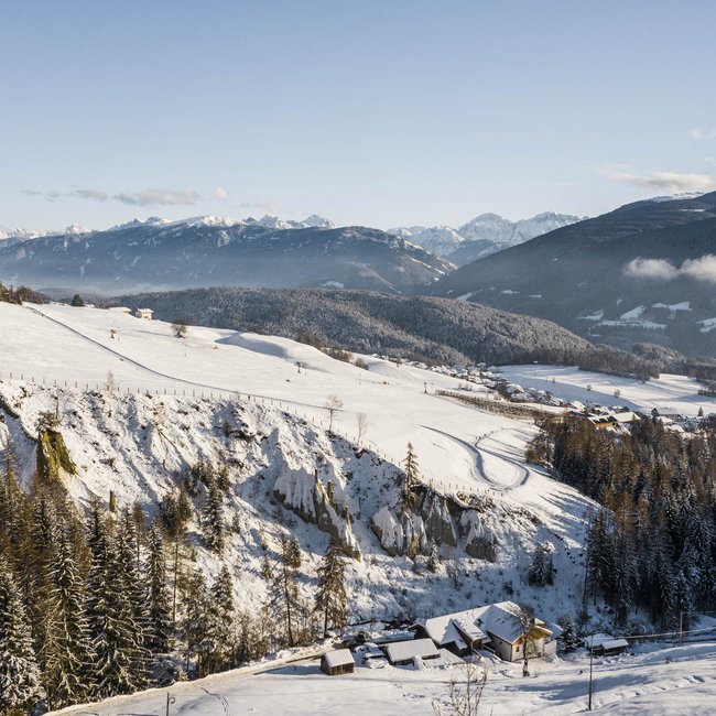 Rodeln in Südtirol Schneebedeckte Alpenlandschaft mit Häusern und Wald unter blauem Himmel