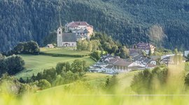 Mächtig und erhaben: Schloss Rodenegg Das Bild zeigt eine Kirche und ein Gebäude auf einem Hügel, umgeben von grünen Wiesen und dichten Wäldern. Im Vordergrund ist unscharfes Gras zu sehen.