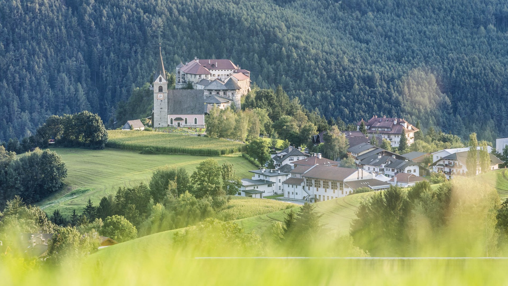 Rodeneck: idylický turistický ráj Obrázek ukazuje kostel a budovu na kopci, obklopené zelenými loukami a hustými lesy. V popředí je rozostřená tráva.