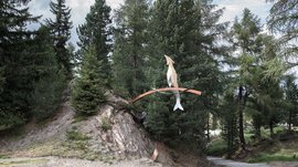 Der Schöpfungsweg in Rodeneck Das Bild zeigt eine Kunstinstallation in einem Waldgebiet. Die Skulptur besteht aus einem abstrahierten Fisch und einem Vogel, die auf einem geschwungenen Metallbogen balancieren