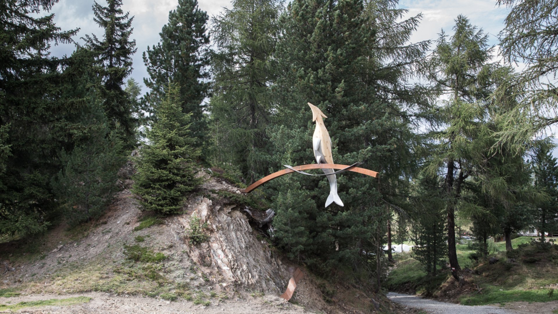 Der Schöpfungsweg in Rodeneck Das Bild zeigt eine Kunstinstallation in einem Waldgebiet. Die Skulptur besteht aus einem abstrahierten Fisch und einem Vogel, die auf einem geschwungenen Metallbogen balancieren