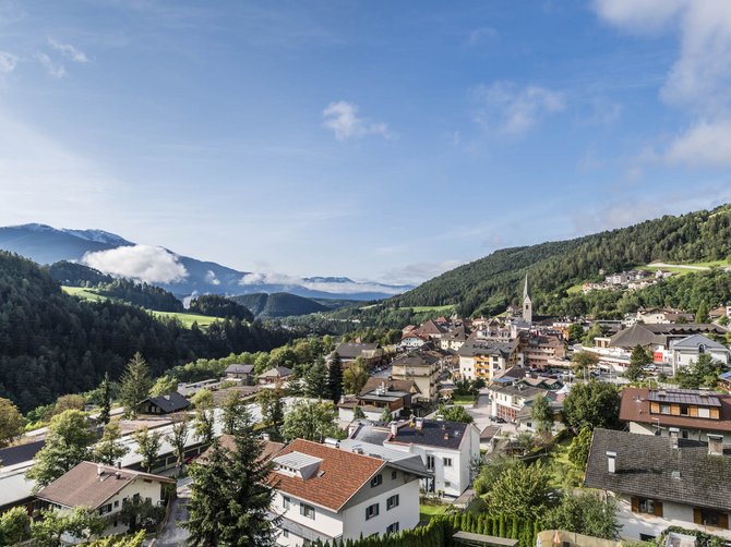 The Alpine hamlet of Spinges The picture shows a picturesque town surrounded by green hills and mountains. In the foreground, houses with red roofs and lush greenery can be seen, while in the background fog-covered mountains and a clear blue sky are visible.