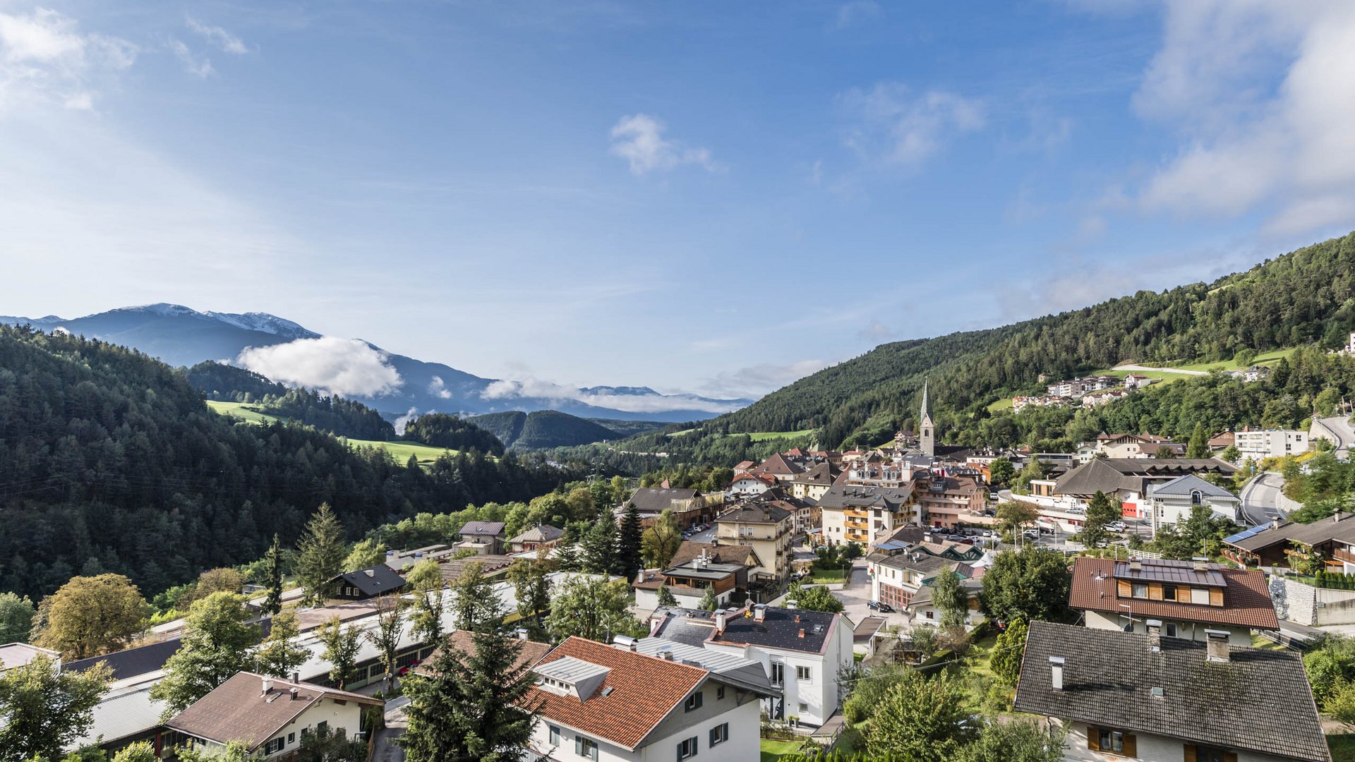 Mühlbach im Pustertal Das Bild zeigt eine malerische Stadt, umgeben von grünen Hügeln und Bergen. Im Vordergrund sind Häuser mit roten Dächern und üppigem Grün zu sehen, während im Hintergrund nebelverhangene Berge und ein klarer blauer Himmel zu erkennen sind.