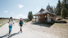 Alpská pastvsina Rodenecker-Lüsner Alm Obrázek ukazuje dva turisty, muže a ženu, kteří kráčejí po široké písečné cestě. Napravo od nich stojí malá kamenná kaple s dřevěnou střechou. Obloha je jasná a modrá a v pozadí jsou vidět mírné kopce a stromy.