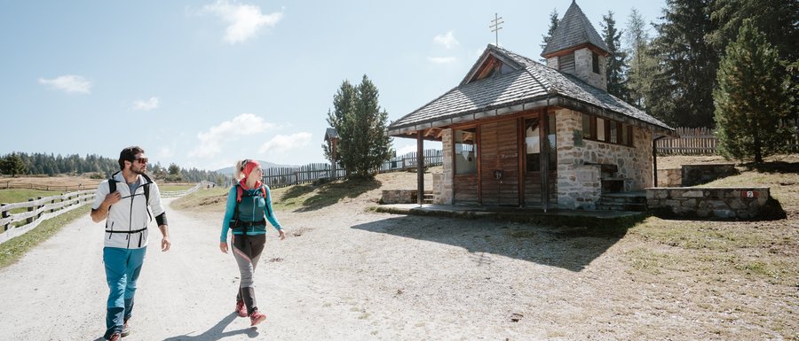 The Gitschberg webcams The picture shows two hikers, a man and a woman, walking along a wide, sandy path. To their right stands a small stone chapel with a wooden roof. The sky is clear and blue, and in the background, gentle hills and trees can be seen.