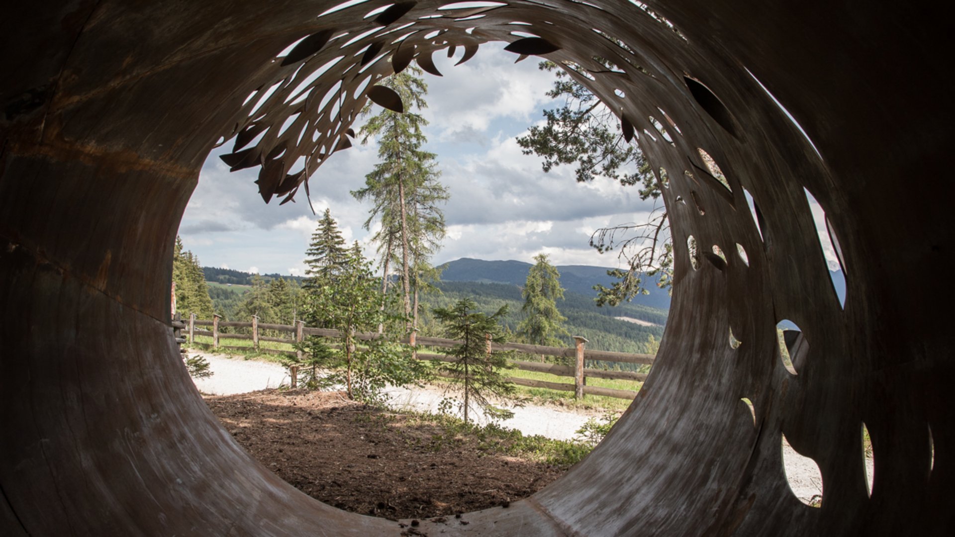 Der Schöpfungsweg in Rodeneck Das Bild zeigt den Blick durch ein großes, rostfarbenes Metallrohr, das kunstvoll mit Blättermustern durchbrochen ist. Draußen sind Bäume, ein Holzzaun und in der Ferne Hügel unter einem bewölkten Himmel zu sehen.