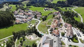 Het Aktivpark Terenten De afbeelding toont een luchtfoto van een klein dorpje dat zich bevindt in een groen heuvelachtig landschap. Te zien zijn talrijke huizen met rode daken, een kerk met een scherpe toren, evenals een grote speeltuin en een sportveld, omgeven door weiden en bossen.