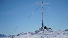 Vals - Jochtal - Steinermandl The picture shows a snow-covered mountain peak with a large summit cross partially covered with ice. Two people in winter clothing stand next to the cross and look into the distance under a clear blue sky.