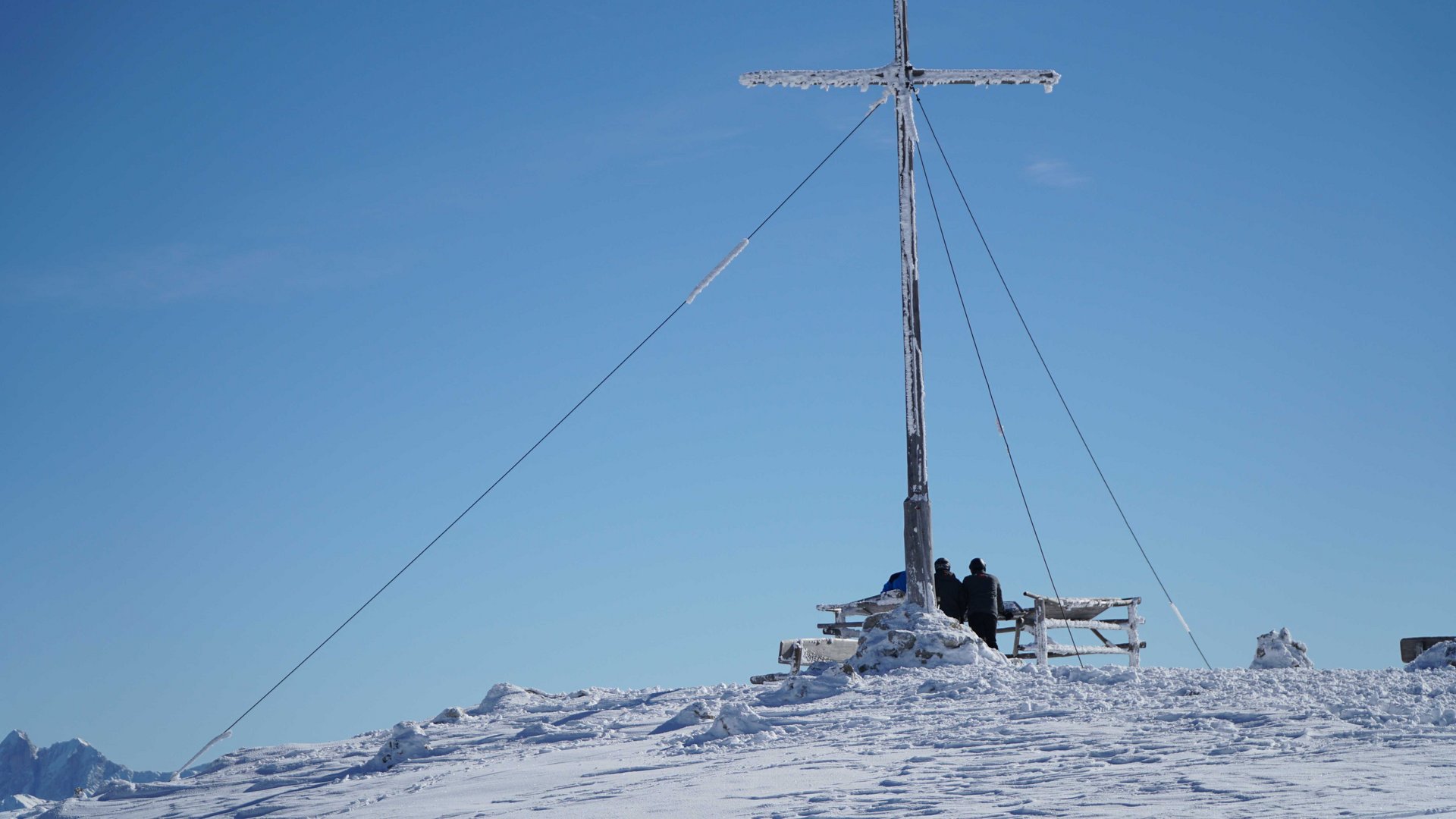 Vals - Jochtal - Steinermandl The picture shows a snow-covered mountain peak with a large summit cross partially covered with ice. Two people in winter clothing stand next to the cross and look into the distance under a clear blue sky.