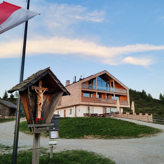 Starkenfeldhütte Obrázek ukazuje moderní třípatrový horský dům s velkými okny a dřevěným obkladem, který stojí na kopci. V popředí vlaje červeno-bílá vlajka vedle dřevěného kříže a obloha je mírně zatažená s jemným večerním světlem.