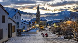 Advent in Terenten The picture shows a snowy village street with a church in the background and festively decorated houses. A family is walking in a picturesque winter landscape at sunset.