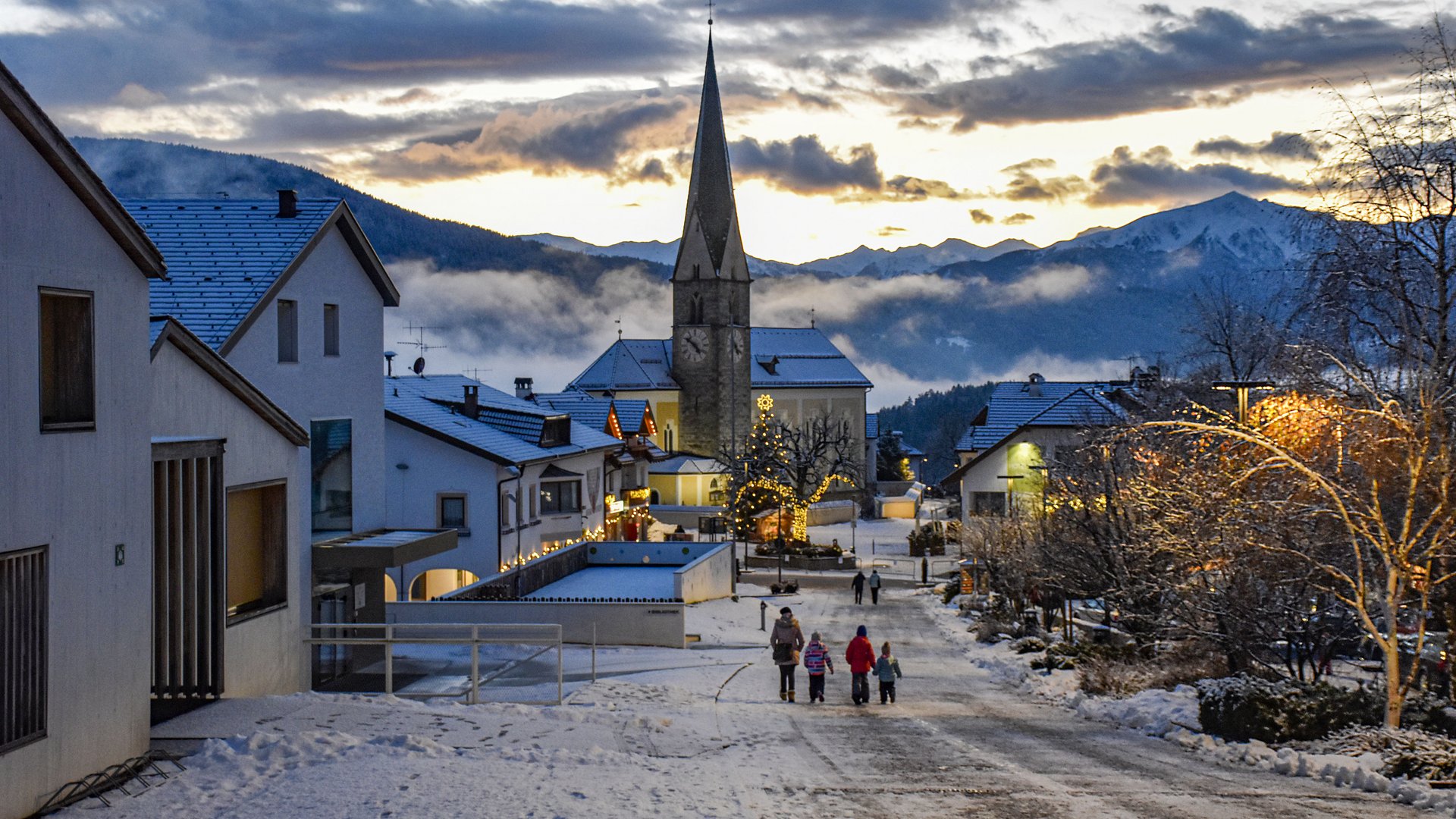 Advent in Terenten The picture shows a snowy village street with a church in the background and festively decorated houses. A family is walking in a picturesque winter landscape at sunset.