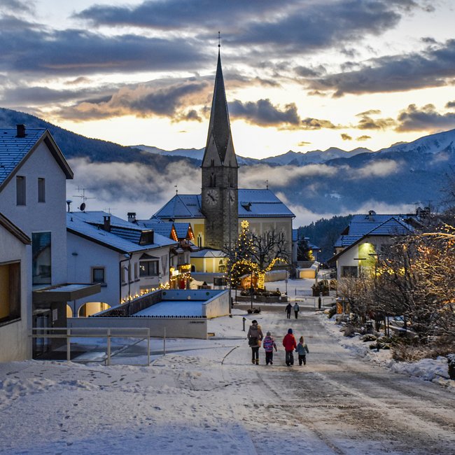 Advent in Terenten The picture shows a snowy village street with a church in the background and festively decorated houses. A family is walking in a picturesque winter landscape at sunset.