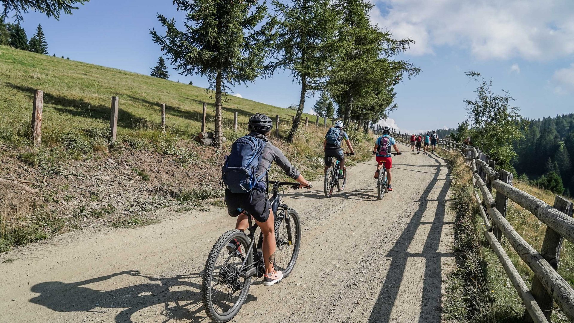 Alpská pastvsina Rodenecker-Lüsner Alm Obrázek ukazuje skupinu cyklistů, kteří jedou do kopce širokou, nezpevněnou cestou. Cesta je lemována stromy, vlevo se rozprostírá zelená louka, zatímco vpravo podél cesty vede dřevěný plot. Krajina naznačuje horské prostředí.