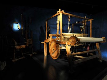Vintl, in the Pustertal valley The picture shows a large wooden loom in a dark room, illuminated by a beam of light. Other historical tools can be seen on the wall in the background.