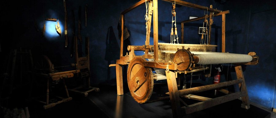 The Gitschberg webcams The image shows a large wooden loom in a dark room, illuminated by a beam of light. In the background, other historical tools can be seen on the wall.
