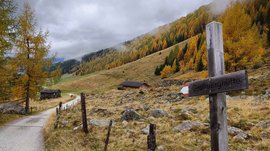 The Altfasstal valley, Meransen The picture shows a hiking trail that leads through an autumnal landscape with colorful, partly yellow and orange trees. The sky is cloudy, and at the roadside stands a wooden signpost with the inscription "Großberghütte." In the background, a few huts and a wooded hill are visible.