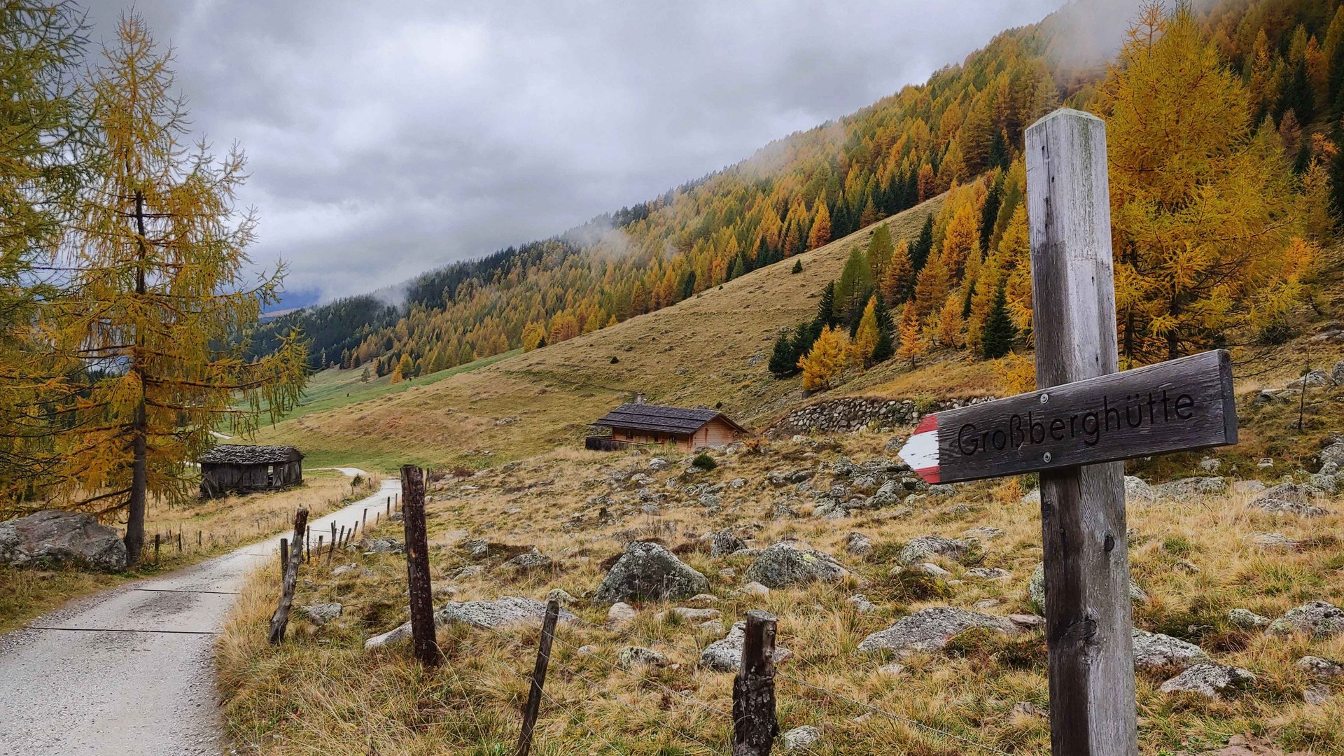 The Altfasstal valley, Meransen The picture shows a hiking trail that leads through an autumnal landscape with colorful, partly yellow and orange trees. The sky is cloudy, and at the roadside stands a wooden signpost with the inscription "Großberghütte." In the background, a few huts and a wooded hill are visible.