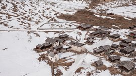 Fane Alm – South Tyrol’s most idyllic Alpine village The picture shows a small village in a snowy mountainous landscape. The houses mostly have wooden or stone roofs, and the surrounding hills are partly covered with snow, while brown areas are visible.