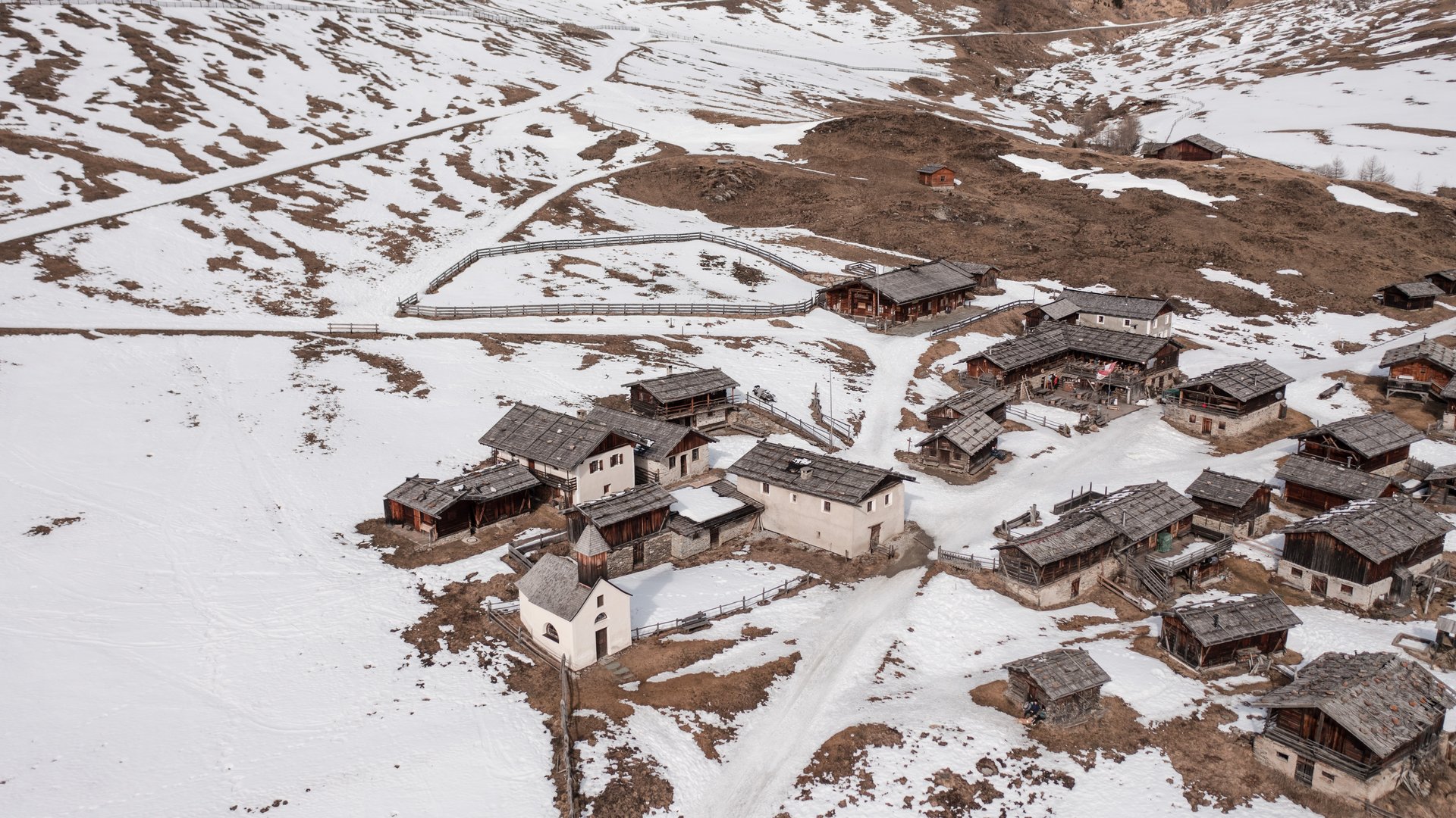 Fane Alm – South Tyrol’s most idyllic Alpine village The picture shows a small village in a snowy mountainous landscape. The houses mostly have wooden or stone roofs, and the surrounding hills are partly covered with snow, while brown areas are visible.