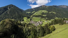 Pfunders: urig und idyllisch Das Bild zeigt ein grünes, hügeliges Tal mit einem kleinen Dorf in der Mitte, umgeben von dichten Wäldern und hohen Bergen. Die Szenerie ist ruhig und idyllisch unter einem strahlend blauen Himmel mit ein paar weißen Wolken.