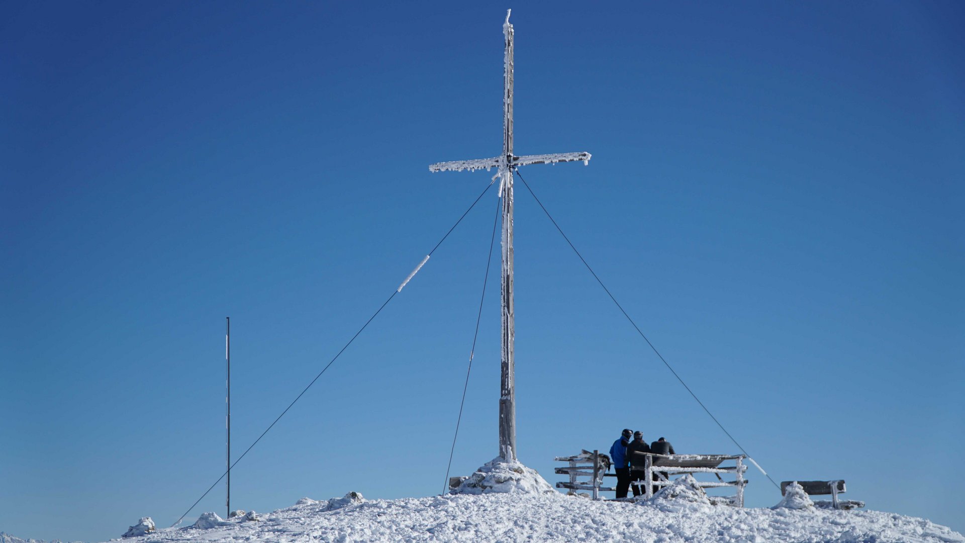 Vals - Jochtal - Steinermandl The picture shows a large summit cross covered with snow and ice, under a bright blue sky. In the background, people stand next to a bench, enjoying the winter view from the mountain peak.